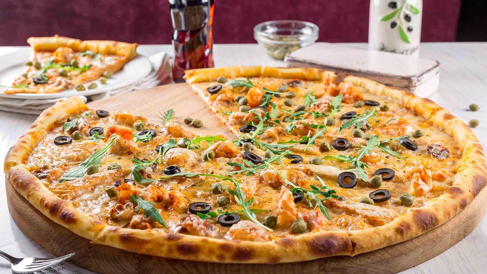 Een verse pizza op een houten plank, met olijven, kappertjes en rucola.